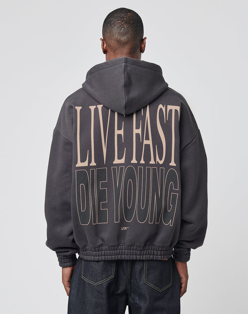 A person wears the LIVE FAST DIE YOUNG Die Young Hooded Zip with an oversized fit and bold beige backprint, featuring front and back prints, a hood, and styled with dark jeans for a relaxed street look.