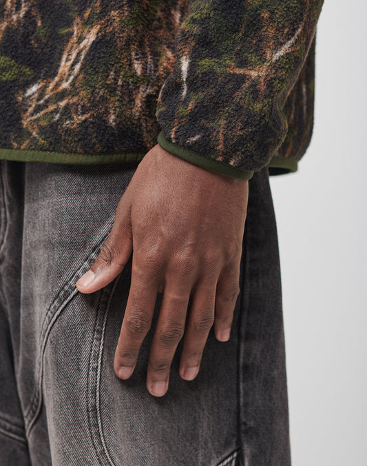 A close-up of a person’s hand at their side, wearing the LIVE FAST DIE YOUNG LFDY Fleece Longsleeve in green and brown polyester with faded gray jeans featuring visible stitching.