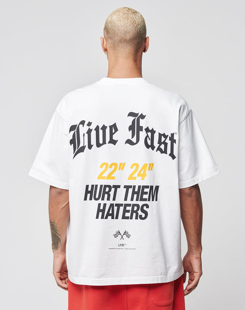 A person with short blond hair, seen from behind, wears the LIVE FAST DIE YOUNG LFDY Haters Tee in oversize fit with bold black text Live Fast and Hurt Them Haters and yellow numbers 22 24. Red shorts finish the outfit.