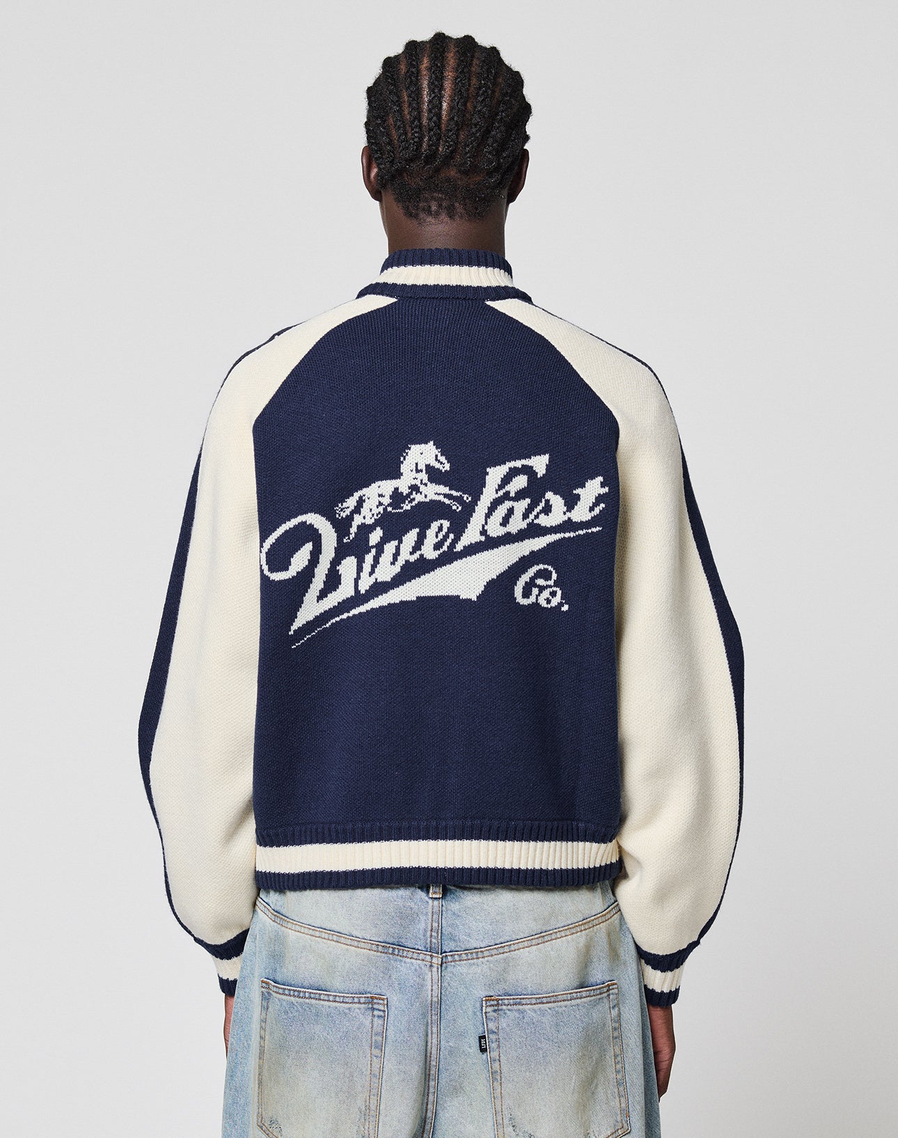 A person with braided hair, seen from behind, wears the Boxy Knit Zip Jacket by LIVE FAST DIE YOUNG with light blue oversize-fit jeans. The navy and cream jacket features a running horse logo and “Live Fast Co.” on the back.