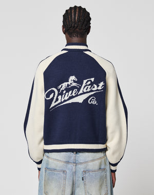 A person with braided hair, seen from behind, wears the Boxy Knit Zip Jacket by LIVE FAST DIE YOUNG with light blue oversize-fit jeans. The navy and cream jacket features a running horse logo and “Live Fast Co.” on the back.