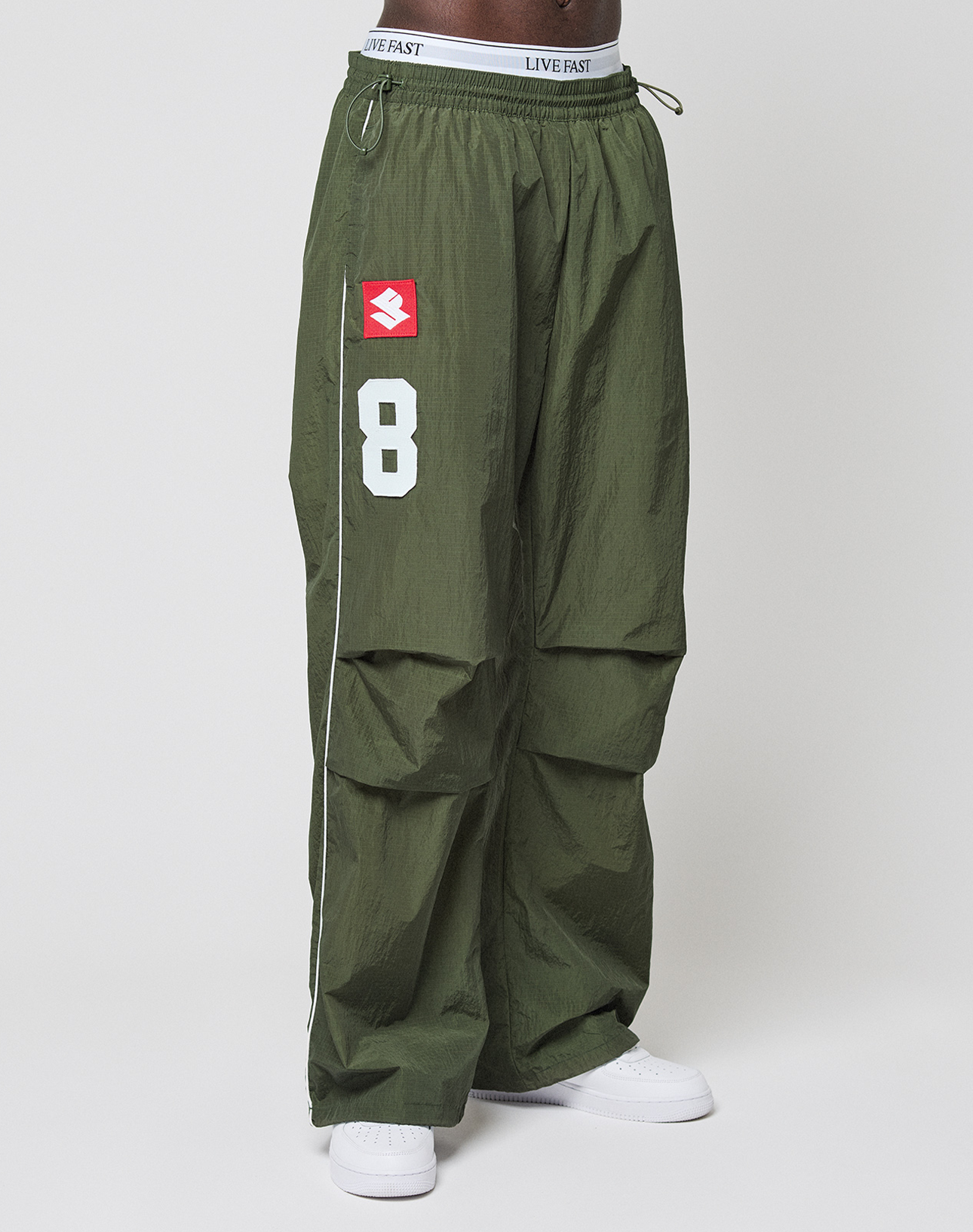 Model wears LIVE FAST DIE YOUNG LFDY Nylon Trackpants in baggy olive green with wide fit, white number 8, red logo-print square, and “LIVE FAST” text on the waistband. Made from polyamide. White sneakers shown on a light backdrop.