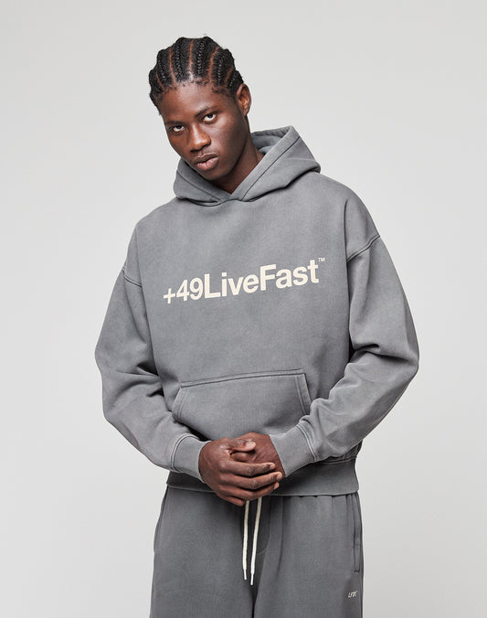 A person with braided hair wears an oversized grey LIVE FAST DIE YOUNG Basic +49 Hooded sweatshirt and matching sweatpants, featuring a bold white +49LiveFast™ frontprint. The background is plain and light-colored.