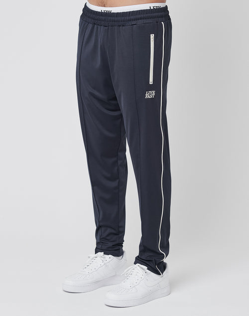 A person wears LIVE FAST DIE YOUNG’s Live Fast Trackjogger in regular fit dark navy with white side piping, logo stitch detail, a vertical zip pocket, and white sneakers, standing against a plain light background; only the lower body is visible.