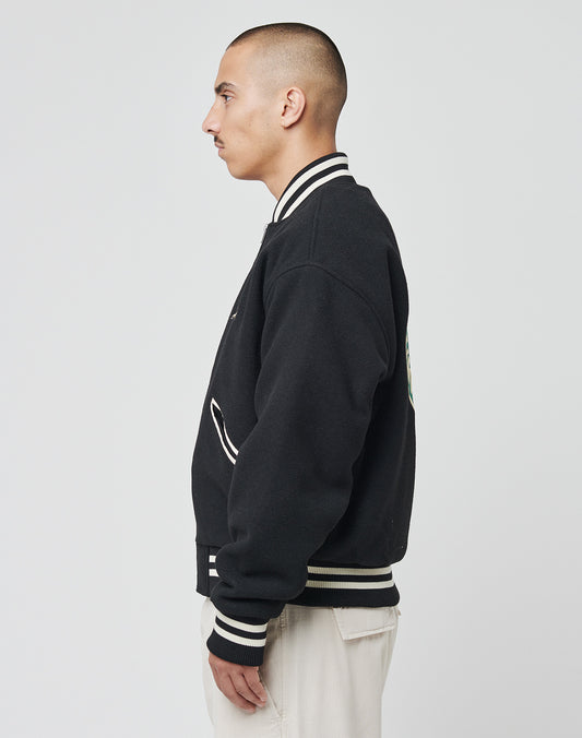 A person with short hair, wearing the LIVE FAST DIE YOUNG Import & Export College Jacket in black with white contrast stripes and light pants, stands in profile against a plain white background.