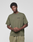 A young man faces the camera, hands loosely clasped, wearing a LIVE FAST DIE YOUNG Basic 210 Tee in olive green with front and back print. The oversize fit and 100% cotton offer comfort and style, paired with matching pants against a plain background.
