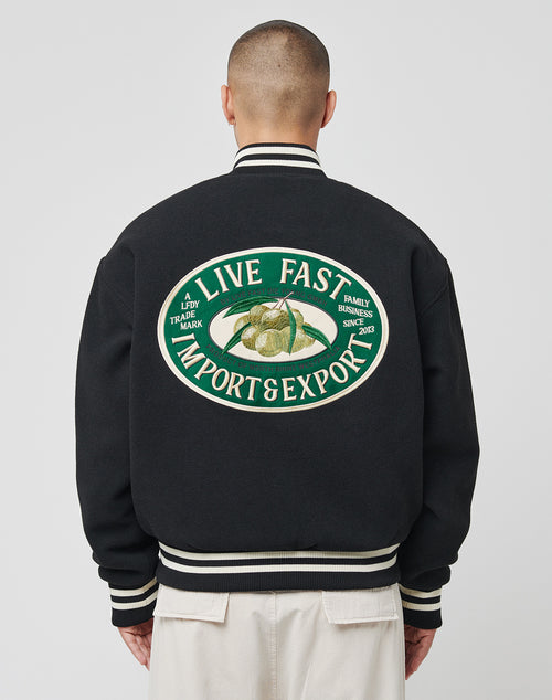 A person seen from behind wears the LIVE FAST DIE YOUNG Import & Export College Jacket—a black polyester varsity with cream accents and a large back logo patch featuring LIVE FAST IMPORT & EXPORT plus durian images and extra text.