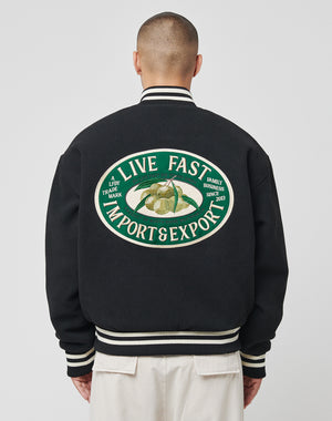 A person seen from behind wears the LIVE FAST DIE YOUNG Import & Export College Jacket—a black polyester varsity with cream accents and a large back logo patch featuring LIVE FAST IMPORT & EXPORT plus durian images and extra text.