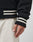 A person wears the LIVE FAST DIE YOUNG Import & Export College Jacket—black polyester with white and black Kontrastfarbe striped cuffs and hem—paired with light pants. The photo focuses on their hand, sleeve, and waistband details.