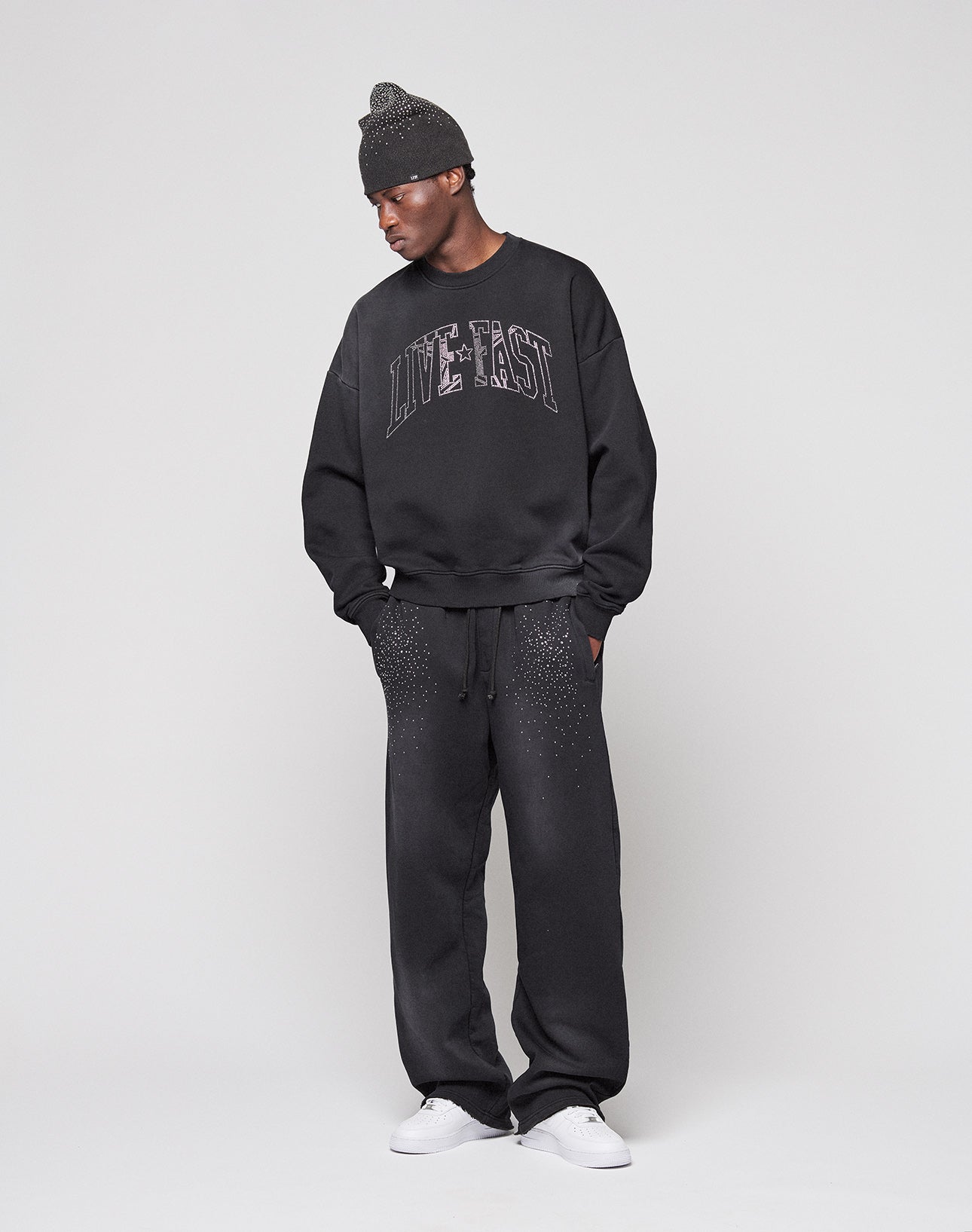 A person in a black beanie, LIVE FAST DIE YOUNG's Live Fast Rhinestone Sweater (oversized fit), black sweatpants with white specks, and white sneakers stands against a plain light background.