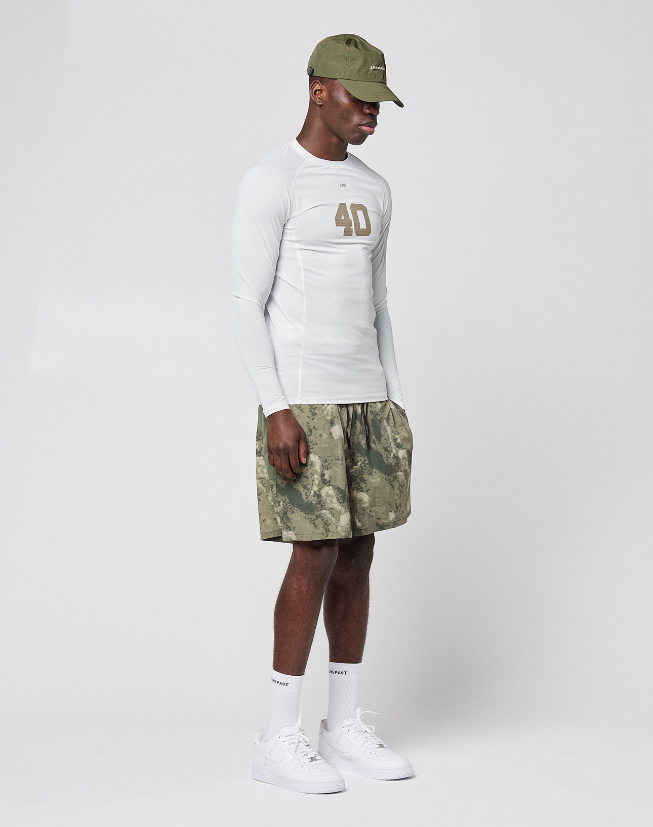 A man wearing the LIVE FAST DIE YOUNG LFDY Performance Slim Longsleeve, with "40" on the front, pairs it with camouflage shorts, white socks and sneakers, and a green cap, standing against a plain white background.