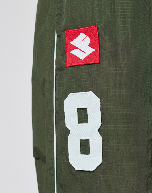Close-up of olive green LFDY Nylon Trackpants by LIVE FAST DIE YOUNG, featuring a sewn white number 8 patch and red patch with a white S logo. A thin white stripe runs vertically beside the patches.
