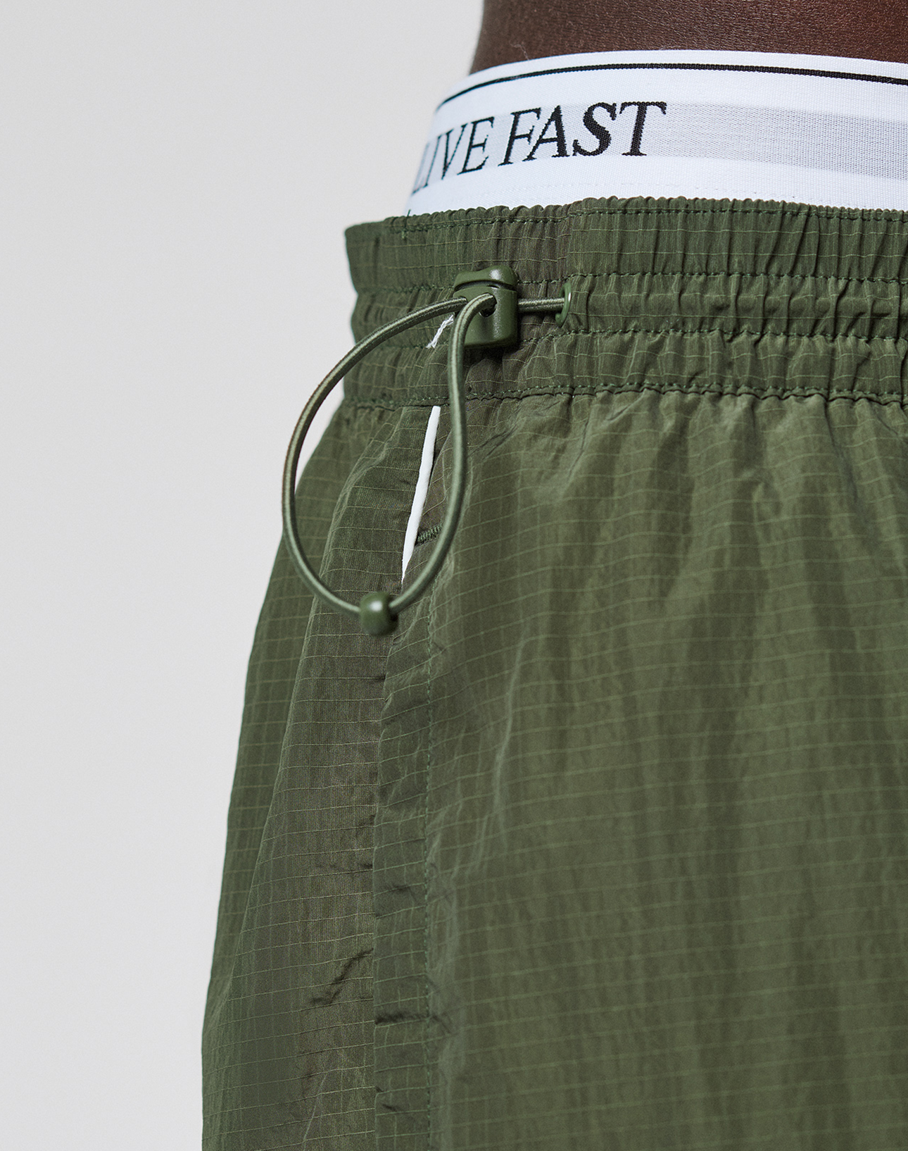 Close-up of LIVE FAST DIE YOUNG’s olive green LFDY Nylon Trackpants featuring a logo print, white waistband with “LIVE FAST” in black, wide fit, cinched elastic waist, polyamide blend fabric, and a drawstring with plastic toggle.