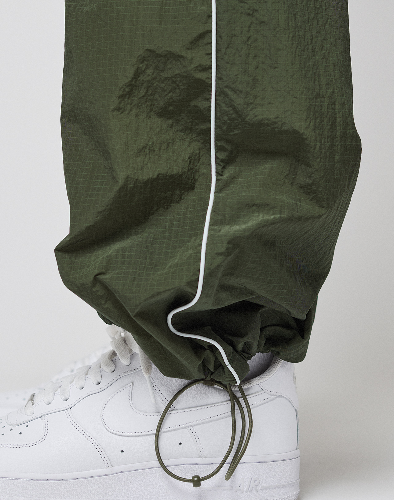 A close-up of a person wearing LIVE FAST DIE YOUNG LFDY Nylon Trackpants in olive green with white piping and a subtle logo print, paired with white Nike Air Force 1 sneakers against a plain background.