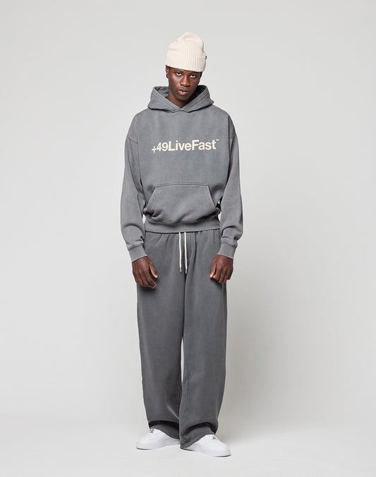 A person wears the Basic +49 Hooded by LIVE FAST DIE YOUNG—an oversized gray hoodie with a front print—paired with matching sweatpants, white sneakers, and a cream knit beanie, all in soft Baumwolle, against a light gray background.
