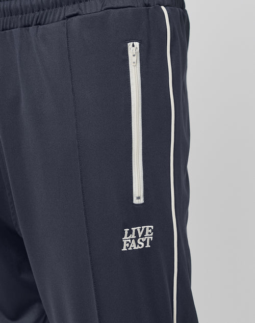 Close-up of the navy blue Live Fast Trackjogger by LIVE FAST DIE YOUNG in Regular Fit, with a vertical white zippered pocket, white piping, and LIVE FAST embroidered below the pocket for a sporty logo detail.