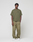 A man with short curly hair and a relaxed pose stands against a plain background in a LIVE FAST DIE YOUNG Basic 210 Tee, olive green and oversize fit, paired with matching loose sweatpants with drawstring and white sneakers.
