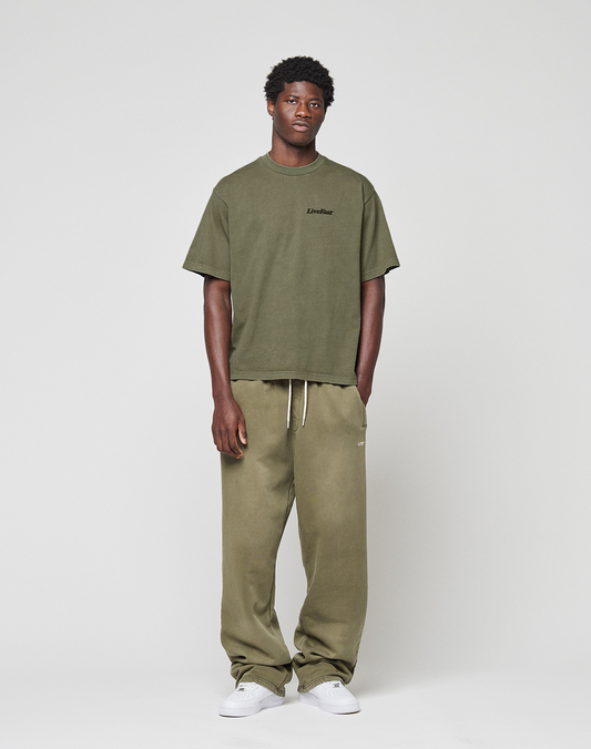 A man with short curly hair and a relaxed pose stands against a plain background in a LIVE FAST DIE YOUNG Basic 210 Tee, olive green and oversize fit, paired with matching loose sweatpants with drawstring and white sneakers.