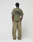 A person faces away, wearing a LIVE FAST DIE YOUNG Basic 210 Tee in olive green with black front and back print, paired with loose matching pants. The background is plain and light-colored.