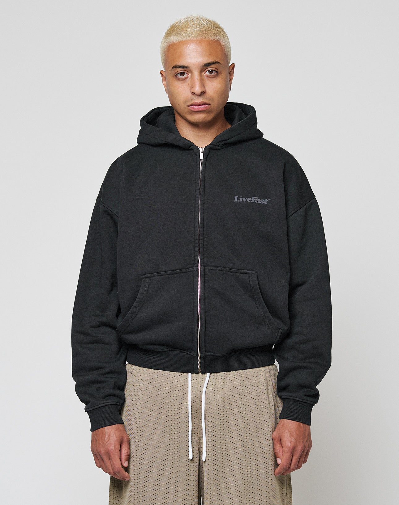 A person with short bleached blonde hair wears the LIVE FAST DIE YOUNG Basic 510 Hooded Zip in black, featuring LiveBase text on the chest, paired with beige textured pants, and stands against a plain white background.