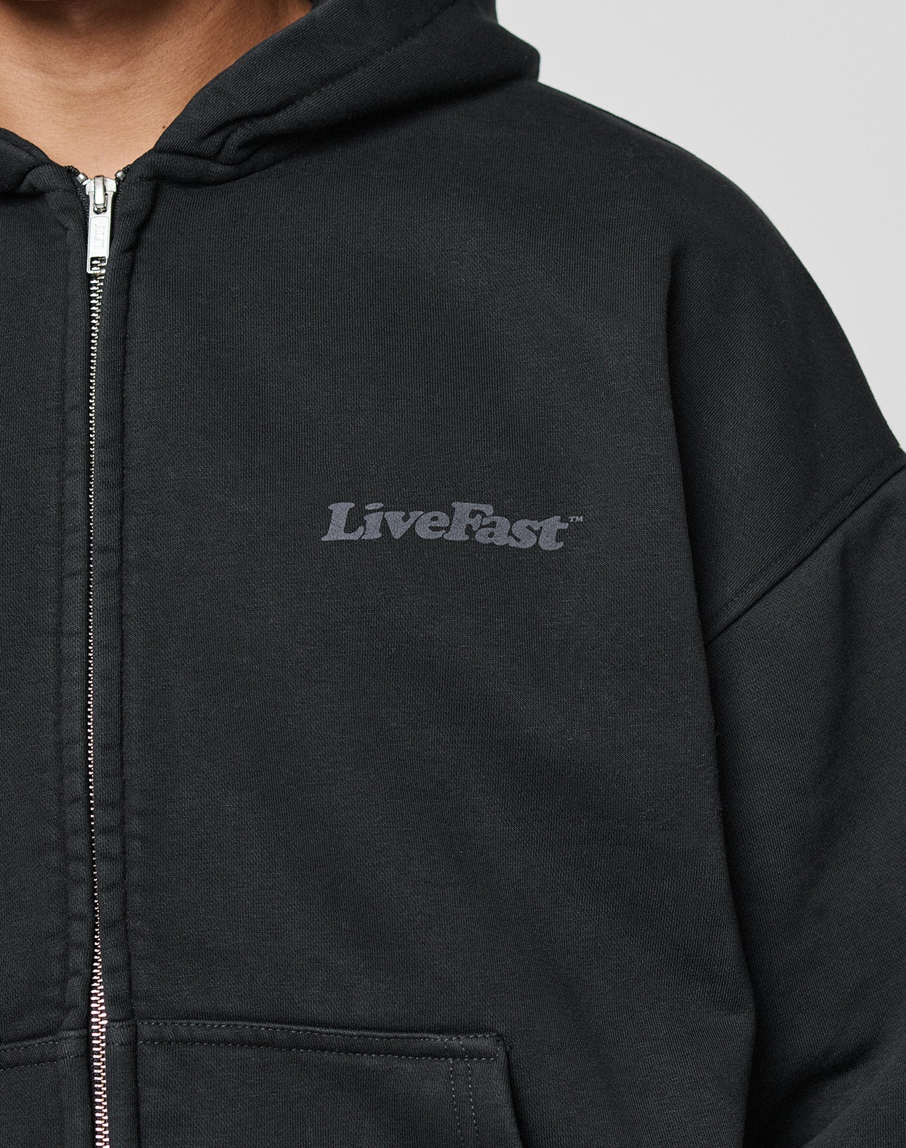 A person wears the LIVE FAST DIE YOUNG Basic 510 Hooded Zip, a black hoodie with LiveFast in gray on the left chest, made from durable 510 gsm fabric. Only their upper torso is shown against a plain background.