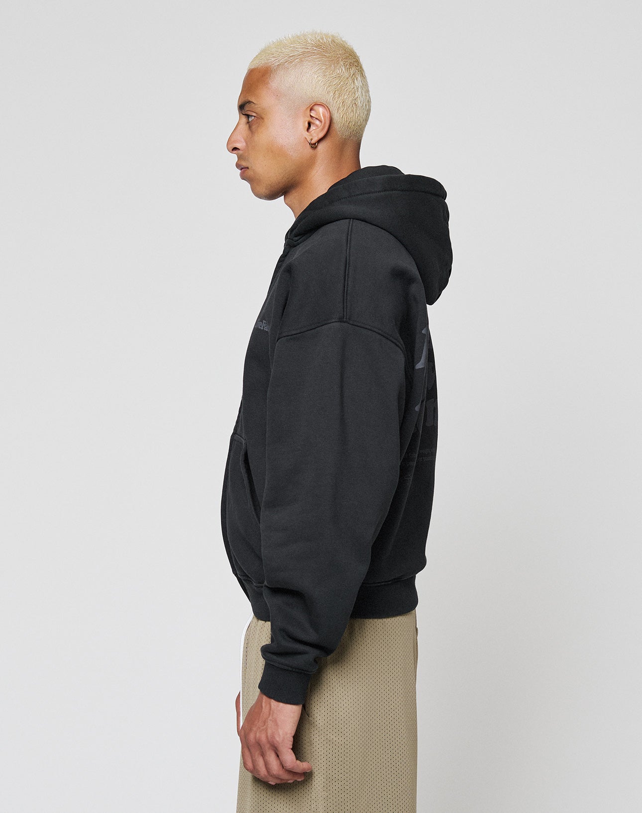 A person with short bleached blonde hair wears the LIVE FAST DIE YOUNG Basic 510 Hooded Zip in black and beige textured pants, standing in profile against a plain light background.