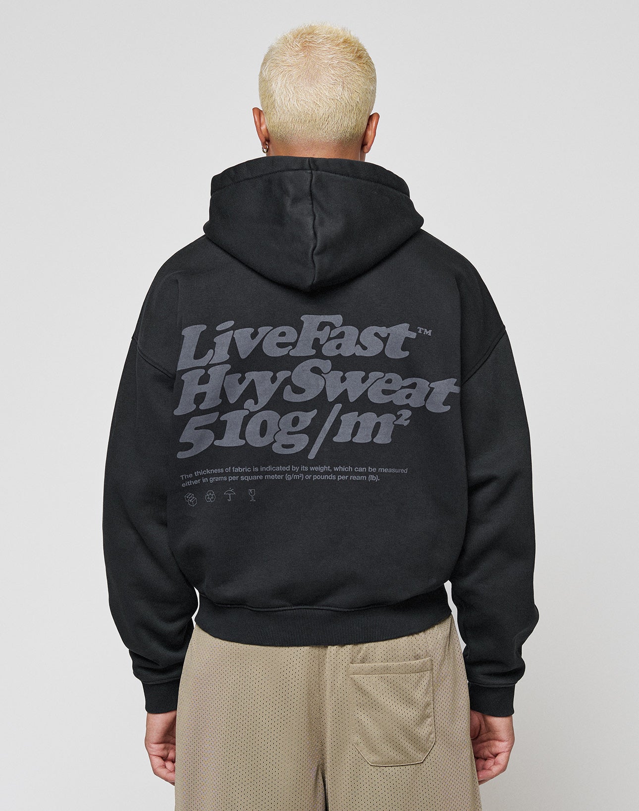 A person with short, bleached blond hair models the LIVE FAST DIE YOUNG Basic 510 Hooded Zip in an oversized fit, featuring bold gray Live Fast, Hvvy Sweat, 510g/m² text on the back. They stand facing away against a plain background.