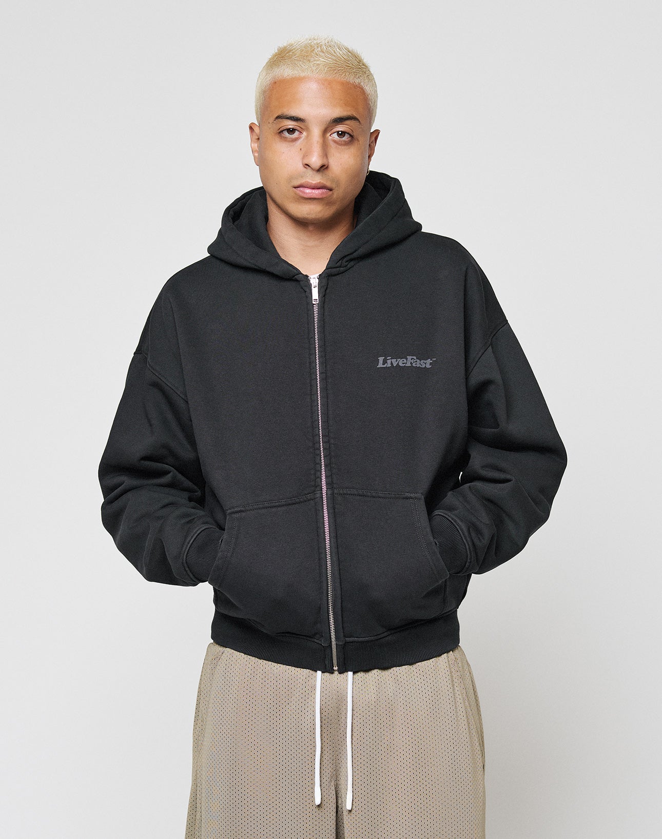A person with short bleached blond hair wears the LIVE FAST DIE YOUNG Basic 510 Hooded Zip in Oversize Fit, featuring LiveFast text on the left chest, paired with light pants, standing against a plain white background.
