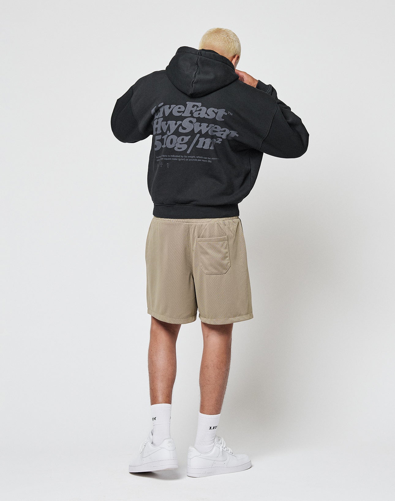 A person with short blond hair faces away, wearing the LIVE FAST DIE YOUNG Basic 510 Hooded Zip in an oversized fit with white text, beige shorts, white socks, and white sneakers, set against a plain white background.