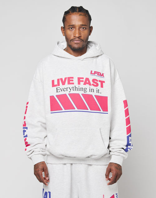 A man wears the LFDY Racing Hooded set by LIVE FAST DIE YOUNG—an oversize fit light gray hoodie and matching sweatpants with bold red and blue glossy LIVE FAST graphics—standing front-facing against a plain background.