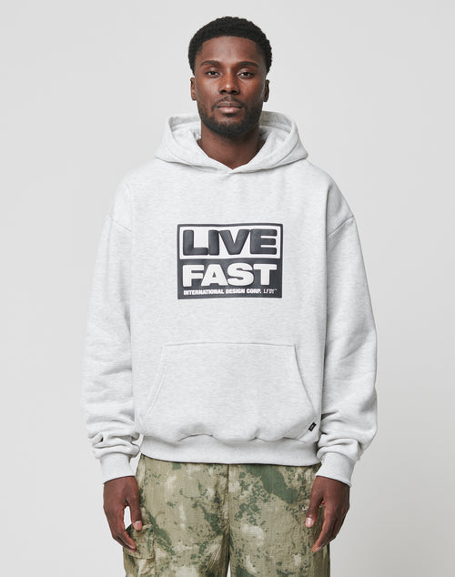A man stands facing forward in an oversized fit, light gray Live Fast Box Hooded sweatshirt by LIVE FAST DIE YOUNG with bold puffer LIVE FAST print on the front, paired with green camouflage pants against a plain light background.