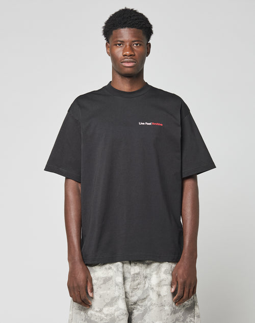 A man stands facing forward against a light gray background, wearing the LIVE FAST DIE YOUNG LFDY F*ck Phones Tee in black with small Live Fast Culture text on the chest and light gray camouflage pants.