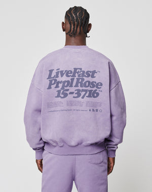 A person with braided hair, seen from behind, wears a LIVE FAST DIE YOUNG Basic Purple Sweater. The vintage-washed sweater features large LiveFast text and PrplRose 15-3716 on the back with additional smaller text below.