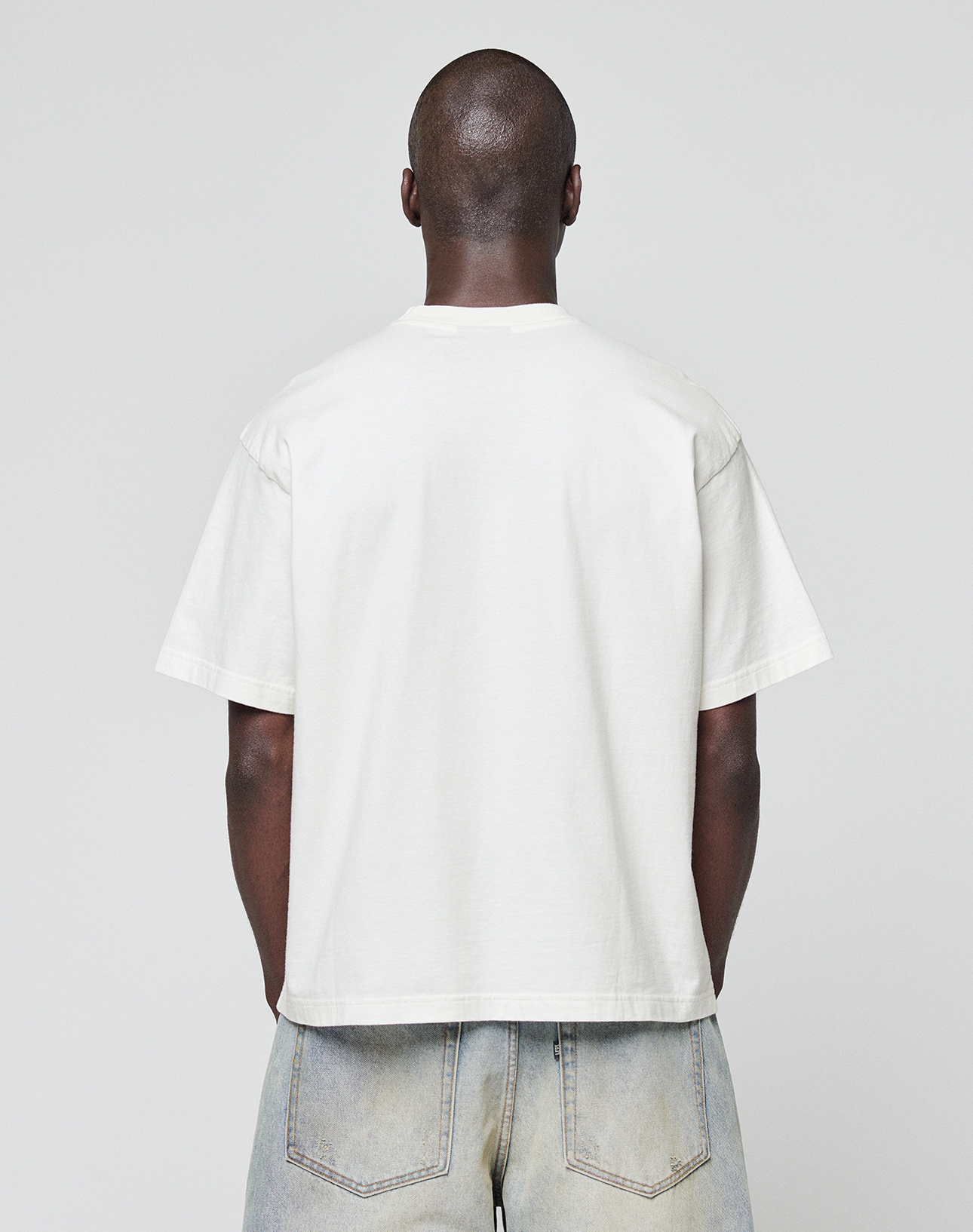 A person with a shaved head stands with their back to the camera, wearing an oversize fit plain white LFDY Block Tee by LIVE FAST DIE YOUNG and light blue jeans against a plain light gray background.