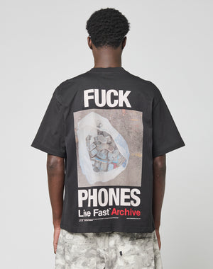 A person is seen from behind wearing the LIVE FAST DIE YOUNG LFDY F*ck Phones Tee, an oversized black cotton shirt with FUCK PHONES and Live Fast Archive plus a bag of old phones graphic on the back. Care instructions are on the label.
