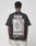 A person is seen from behind wearing the LIVE FAST DIE YOUNG LFDY F*ck Phones Tee, an oversized black cotton shirt with FUCK PHONES and Live Fast Archive plus a bag of old phones graphic on the back. Care instructions are on the label.