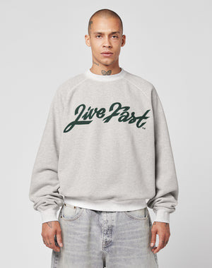 A person with a shaved head models the LIVE FAST DIE YOUNG LFDY Script Sweater—an oversize gray sweatshirt with bold green “Live Fast” text and raglan sleeves—facing forward against a plain light gray background.