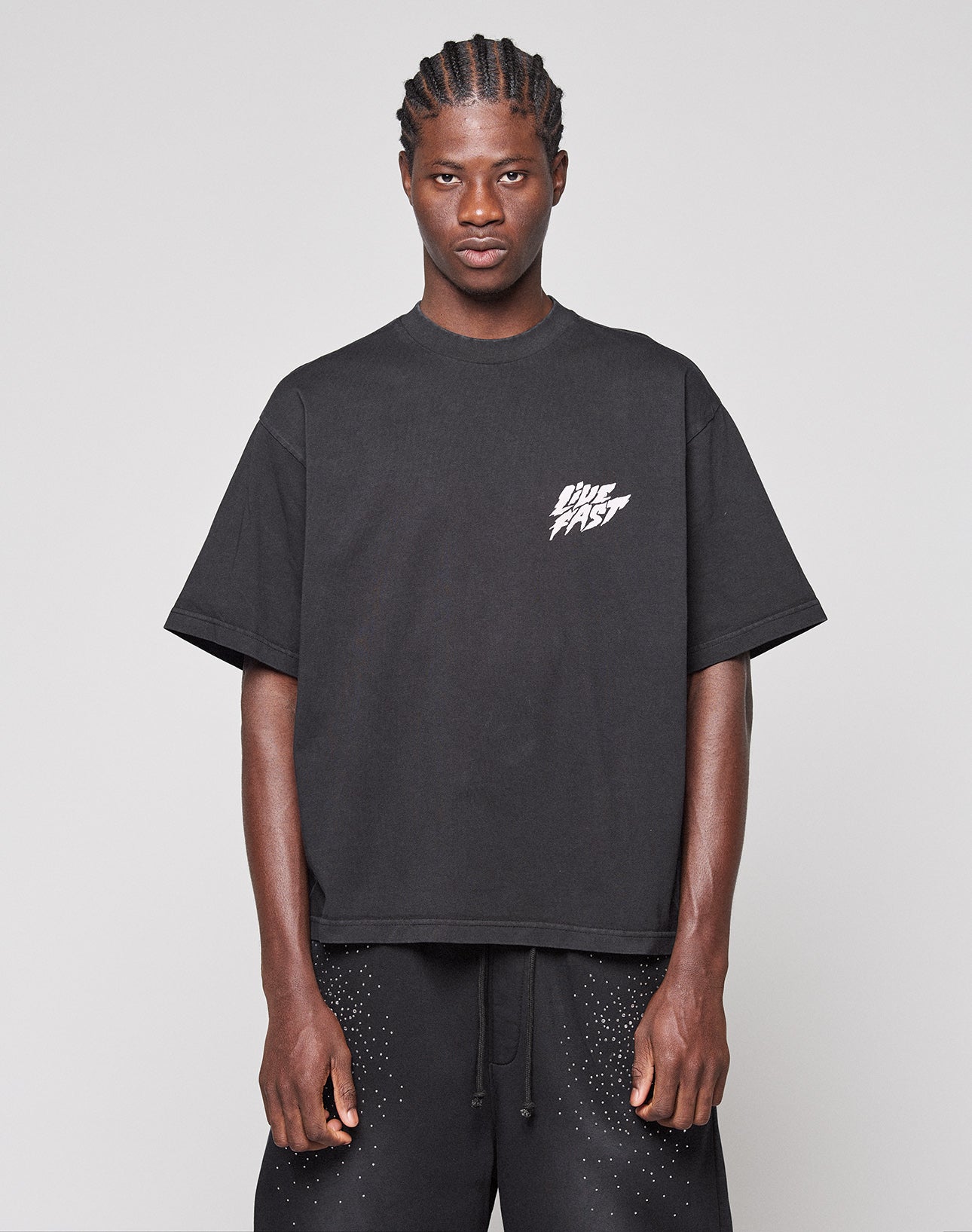 A person stands against a plain background wearing the LIVE FAST DIE YOUNG LFDY Logo Tee, a loose black t-shirt with bold "DIE FAST" frontprint lettering, paired with matching black oversize fit shorts with white speckles.