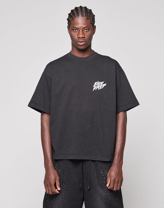 A person stands against a plain background wearing the LIVE FAST DIE YOUNG LFDY Logo Tee, a loose black t-shirt with bold "DIE FAST" frontprint lettering, paired with matching black oversize fit shorts with white speckles.