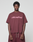 A person wears the Basic +49 Tee by LIVE FAST DIE YOUNG, an oversize maroon shirt with pink +49LiveFast front print, made from 100% Baumwolle, styled with matching maroon pants against a plain light gray background.