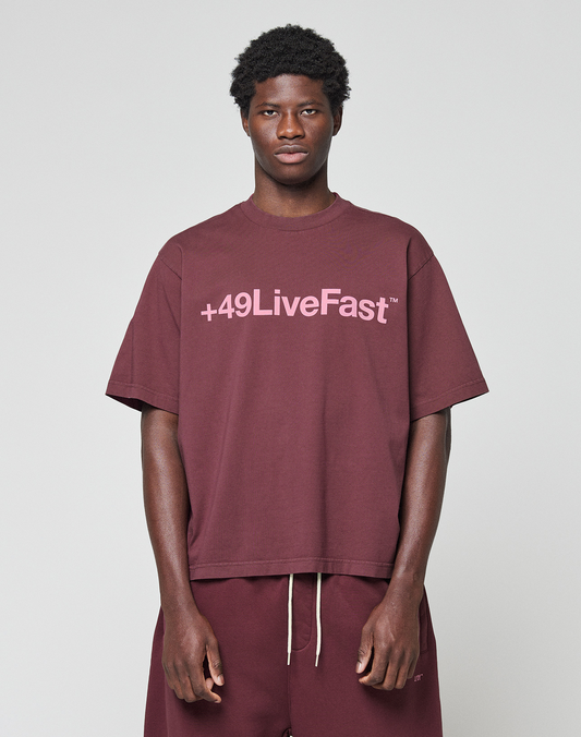 A person wears the Basic +49 Tee by LIVE FAST DIE YOUNG, an oversize maroon shirt with pink +49LiveFast front print, made from 100% Baumwolle, styled with matching maroon pants against a plain light gray background.