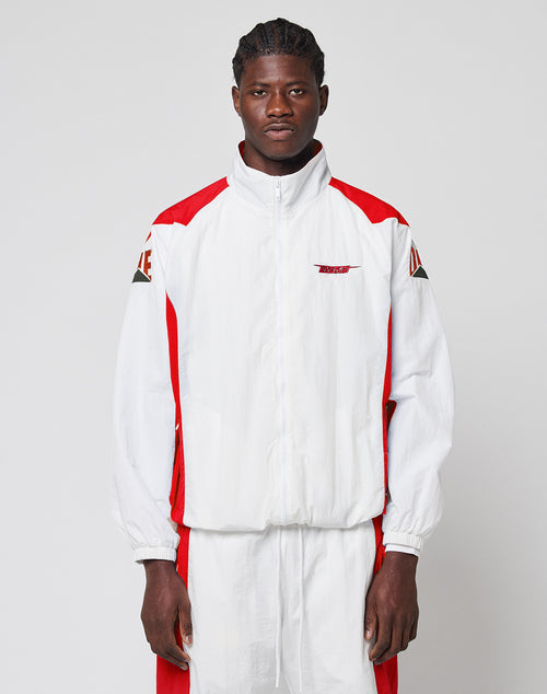 A man stands against a plain background wearing the LFDY Trackjacket 3000 by LIVE FAST DIE YOUNG, a white and red zip-up jacket with branded zipper puller, multi-logo embroidery, red accents, graphic patches, and matching regular fit track pants.