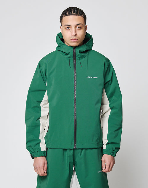 A person faces forward in a green and beige oversize fit Basic Shell Jacket by LIVE FAST DIE YOUNG, featuring a water-resistant zipper and LIVE & START on the chest, paired with matching pants, against a plain white background.
