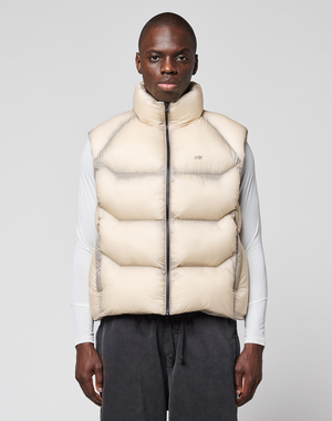 A man faces forward against a light gray background, wearing the LIVE FAST DIE YOUNG Live Fast Puffer Vest in light beige over a white long-sleeve shirt and dark drawstring pants.
