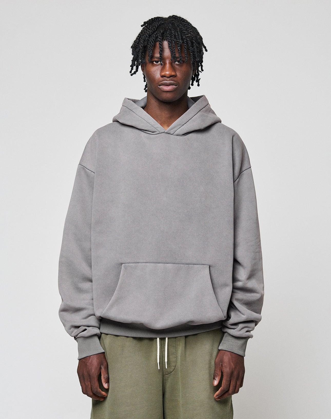 A person with short, twisted hair wears the LIVE FAST DIE YOUNG Basic Blank Hooded in oversize fit and olive green pants, standing against a plain light gray background.