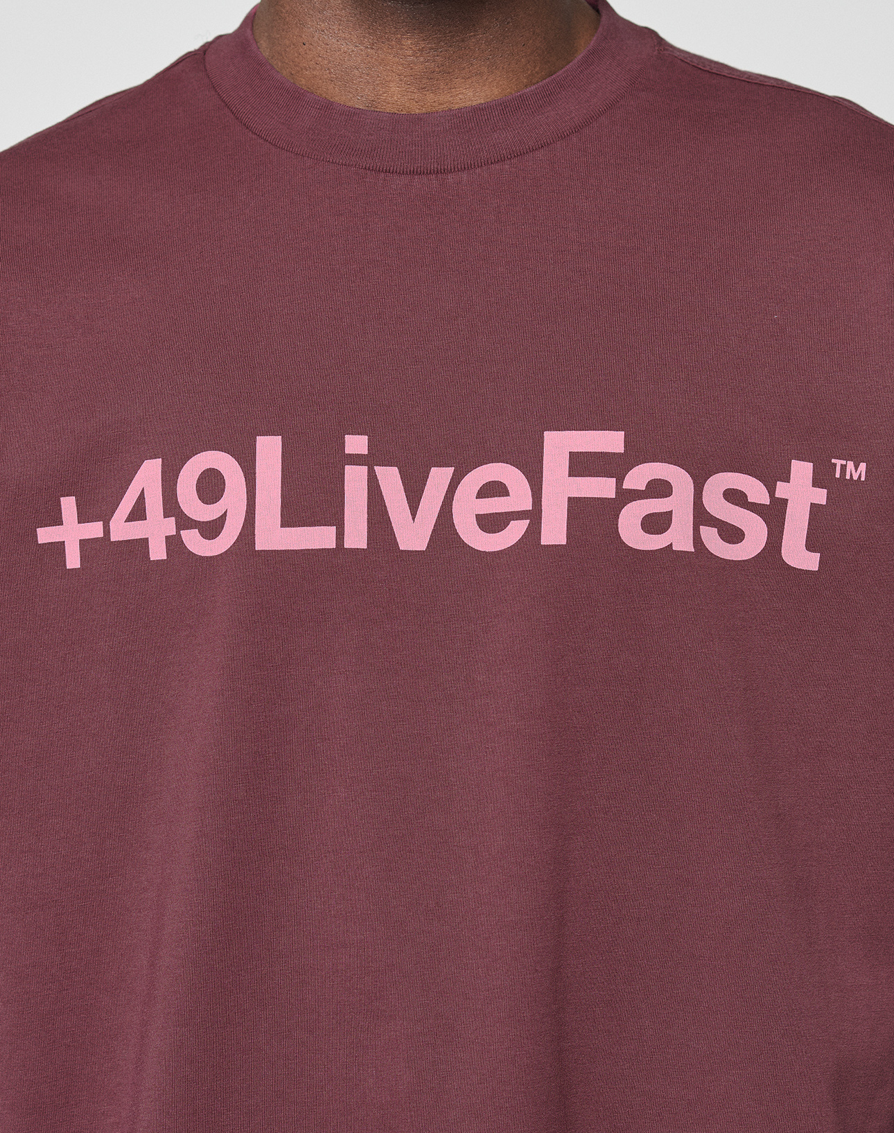 A person wearing the LIVE FAST DIE YOUNG Basic +49 Tee, an oversize fit maroon shirt made from 100% Baumwolle, featuring a pink +49LiveFast™ front print across the chest.