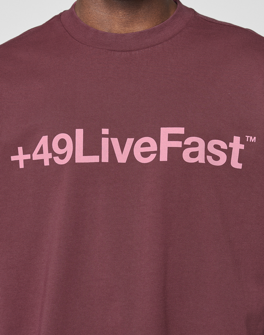 A person wearing the LIVE FAST DIE YOUNG Basic +49 Tee, an oversize fit maroon shirt made from 100% Baumwolle, featuring a pink +49LiveFast™ front print across the chest.
