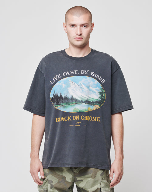 A person with a shaved head wears the Black Chrome Tee by LIVE FAST DIE YOUNG—an oversized 100% Baumwolle T-shirt with mountain and tree prints on front and back, “LIVE FAST, DY. GmbH” above, “BLACK ON CHROME” below—paired with camo shorts.