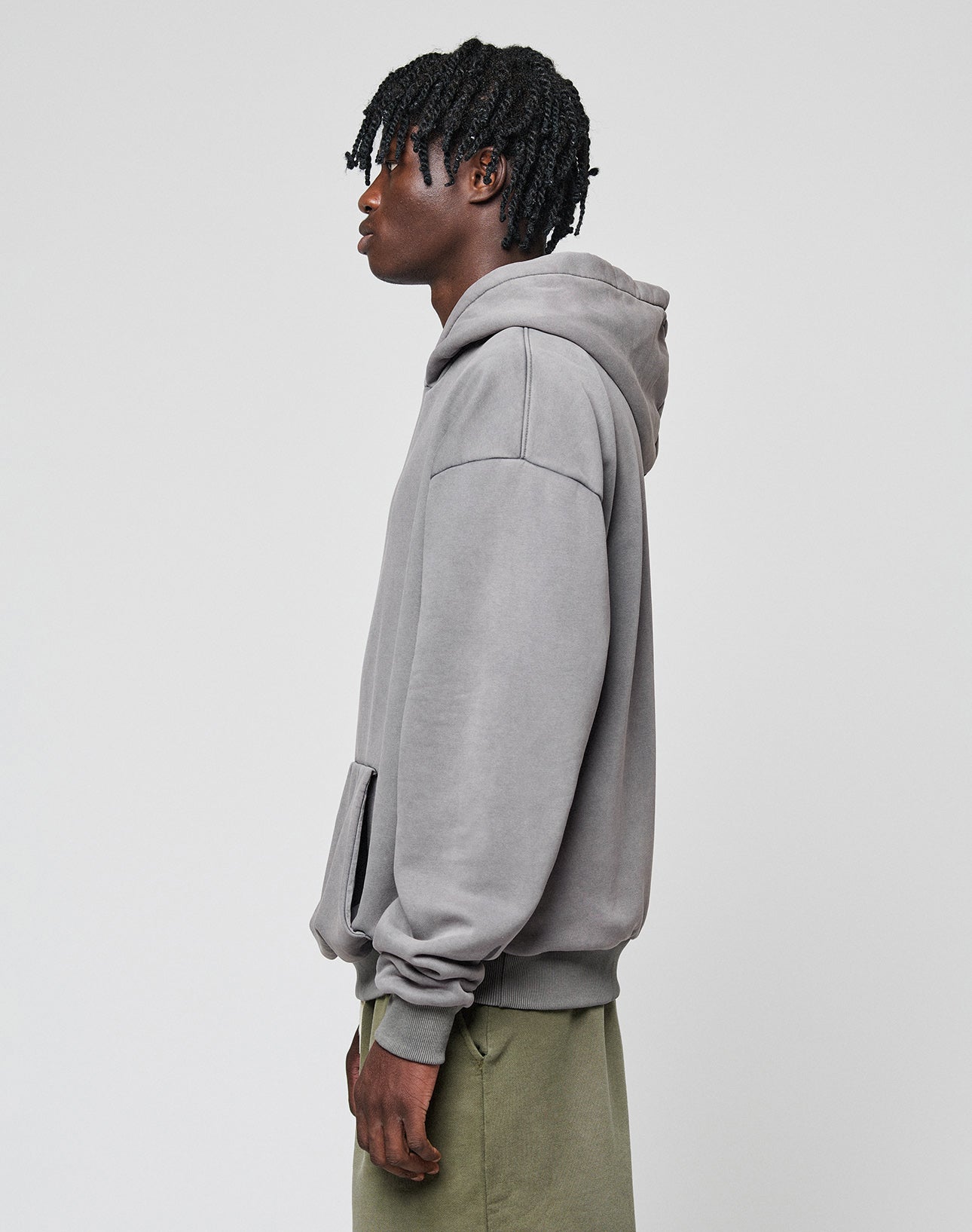 A person with short, textured hair stands sideways in a loose LIVE FAST DIE YOUNG Basic Blank Hooded hoodie and olive green pants against a plain light gray background.