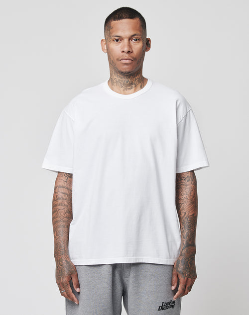 A man with tattoos on his neck, arms, and hands stands facing forward, wearing the LFDY Blank Tee by LIVE FAST DIE YOUNG in white (100% cotton, Regular Fit) and light gray sweatpants against a plain light gray background.
